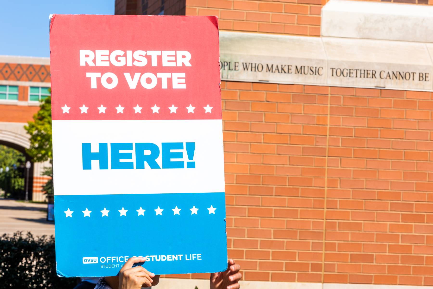 A student holding up a red, white, and blue sign that says "Register to vote here!"
